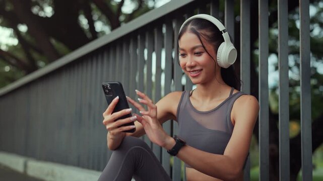 Smiling young Asian woman in sportswear using smartphone and wearing headphones while resting at public park cheerful female tracking workout data on mobile app urban fitness lifestyle concept