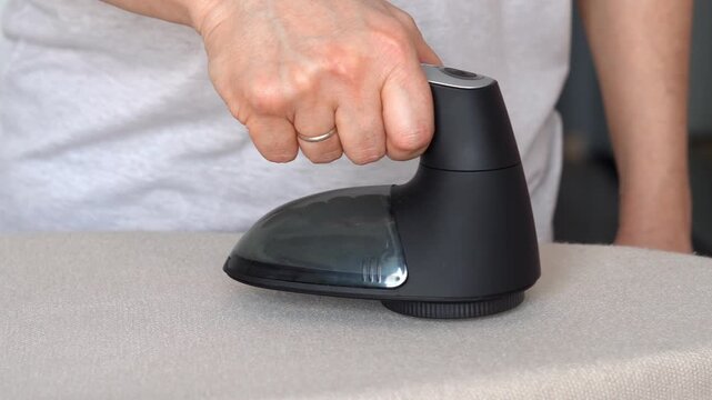 Close up of hand removes pills from knitted clothing using fabric shaver. Textile pilling. Linting. Fabric lint, pilly, pellet, fluff. Fluff-ball remover. Household