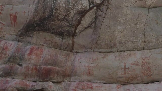 Detailed pictographs on rock face showcasing red and ochre hues, seen beneath a sunlit canopy.