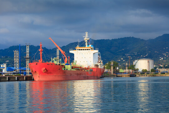 Chemical tanker is moored at industrial port terminal for loading operations. Large chemical tanker features red hull and tall white superstructure set against scenic mountainous landscape background.