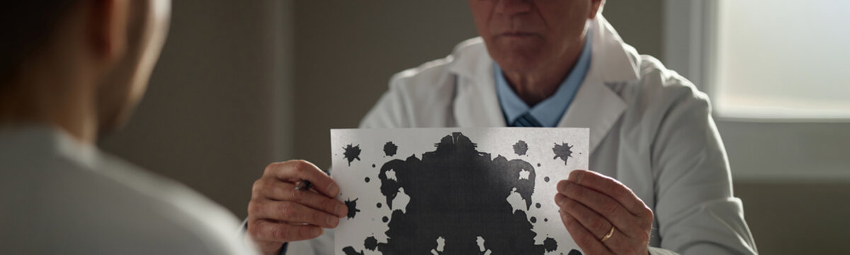 Website header shot of senior male therapist holding inkblot test card during mental health evaluation with patient, assessing emotional state. Useful for psychology, counseling, diagnosis