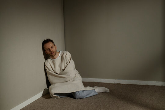 Wide shot of Caucasian young adult man sitting restrained in straitjacket on floor, conveying mental health crisis and isolation. Useful for therapy, psychiatry, awareness campaigns