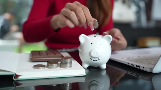 Close-up of a woman putting coins into a white piggy bank, representing saving money, personal finance, budgeting, and future financial security.