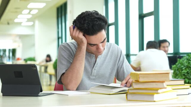 Young Caucasian male student reading a textbook and writing notes on notebook in university library. Teenage guy using a digital tablet and studying for an exam. Education concept.