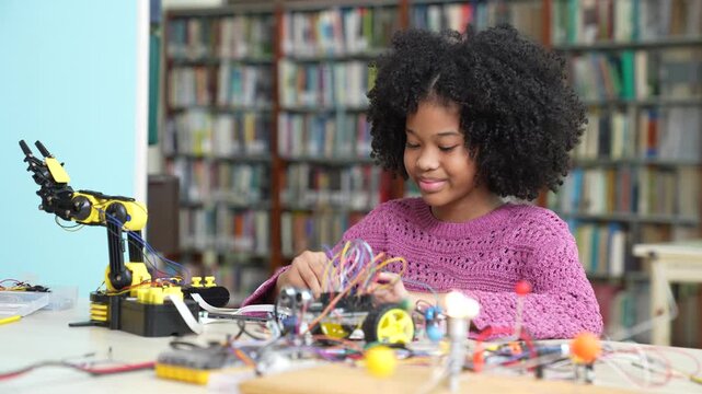 Happy young afro girl with Afro hair building a robot and electronic car in library. Clever African American female student learning STEM education, robotics, engineering project.