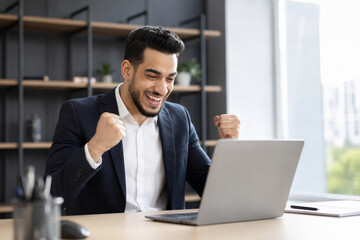 Fototapeta premium Happy businessman in a suit celebrates success while looking at his laptop screen