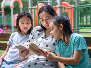 Obraz premium Asian mother reading a book to her two young daughters while sitting on a bench in the park.