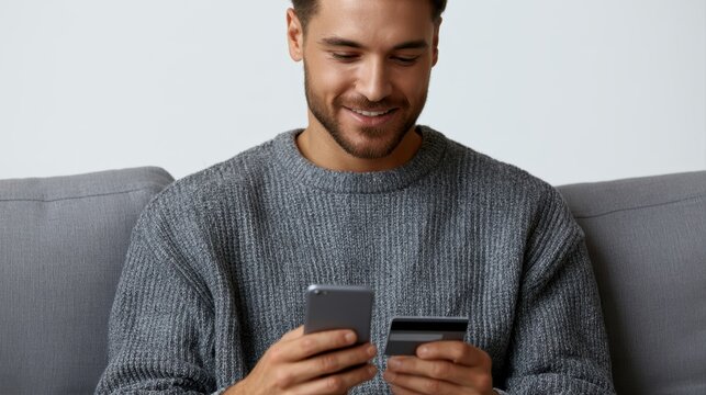 young man smiling at smartphone while holding credit card on couch, online shopping concept