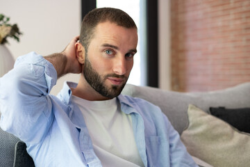 Fototapeta premium Relaxed Young Man With Beard Sitting on Sofa in Modern Living Room Wearing Casual Blue Shirt
