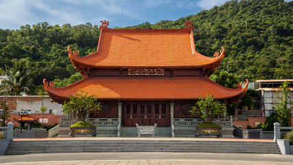Obraz premium Magnificent Ho Chi Minh memorial temple with traditional double-tiered orange tiled roof and ornate wooden carvings, set against lush green hills in Vietnam.