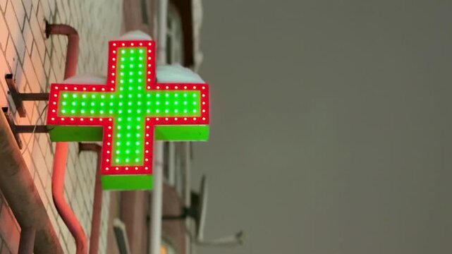 Neon green pharmacy cross signboard mounted on brick facade at dusk, glowing diode lights with red border, metal bracket