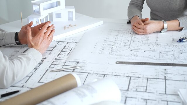 Architects discussing architectural blueprints and a building model on a white desk, planning a new construction project and developing innovative design solutions collaboratively, close up