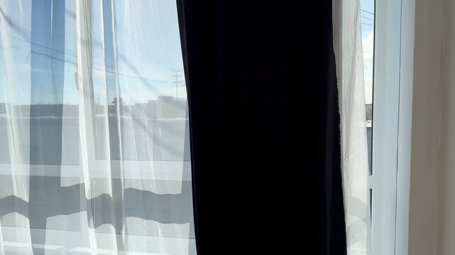 Sunlit window with white tulle drifting across dark curtains, gentle breeze animating sheer fabric, bright blue sky and rooftop