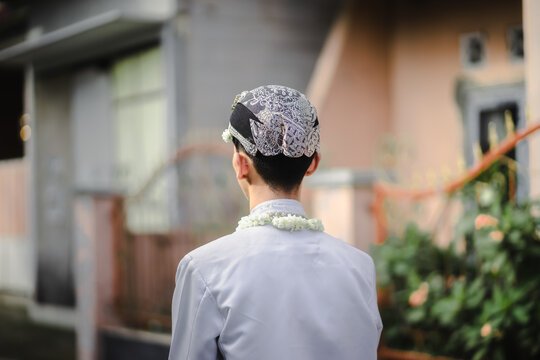 Rear view of a groom wearing an elegant white lace songkok cap and a fresh jasmine flower garland around his neck. Traditional Indonesian wedding attire captured outdoors with soft background blur