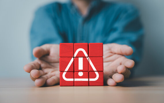 Warning and risk management concept with red alert symbol on wooden blocks protected by human hands. Business security, crisis prevention, safety awareness and problem control concept.