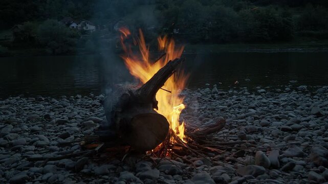 4K video of a campfire positioned slightly left of center and farther back on a stony riverbank at dusk, recorded from Serbia with Bosnian mountains visible across the Drina River.