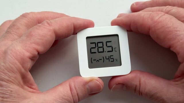 Closeup woman's hands holding digital hygrometer displaying temperature and humidity, small white square sensor, floral