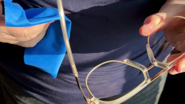 Closeup woman cleaning eyeglasses with blue microfiber cloth, sunlight catching gold frames and hands, slow deliberate wiping