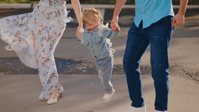 Young family holds hands, skipping joyfully outdoors. Child giggles, eyes bright, feet mid-air with delight. Mom and dad smile, arms linked in playful motion