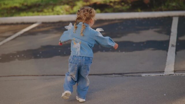 Little girl skips joyfully in parking lot. She wears denim outfit with puffed sleeves. Her arms fling wide as she moves forward. Sunlight glints off pavement behind her