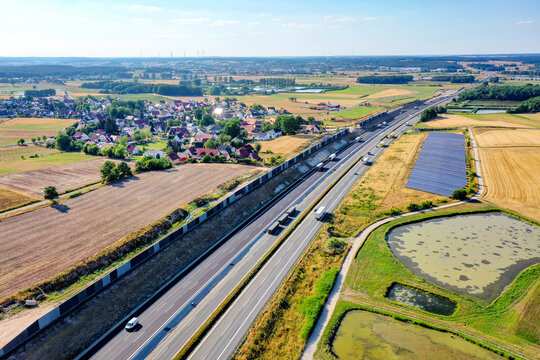 Nurnberg, Germany - 18 May 2024: Aerial view of the A73 highway with traffic, noise barriers, solar panels, and a small village surrounded by green fields and ponds.