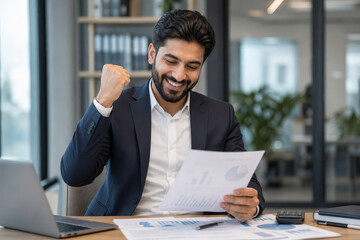 Obraz premium Happy man in a suit celebrates success while holding a document and looking at charts on his desk.