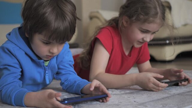 Children Playing Video Games In Mobile Phone at Home Lying on Floor. Mobile phone addiction. Girl and Boy hold smartphone device. Social media obsession. Online communication problem.