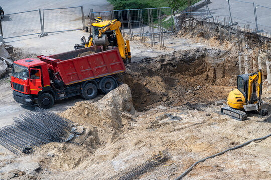 an active construction site where a yellow excavator is loading dirt into a red dump truck next to a deep excavation pit and bundles of steel rebar.