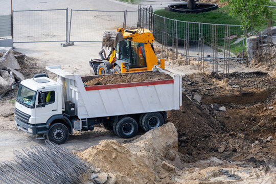 A white dump truck loaded with soil stands near an excavated pit alongside a yellow backhoe loader on an active construction site.