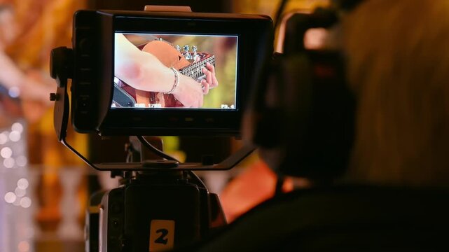 Camera operator filming live acoustic guitar performance on stage using professional broadcasting monitor.