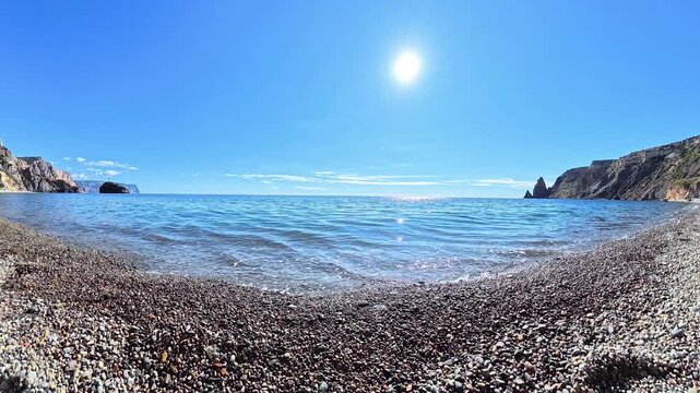 Pebble beach waves gently wash over stone shore under bright summer sun in clear blue sky at scenic rocky coastline