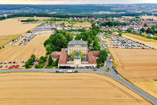 Eggolsheim, Germany - 09 May 2026: Aerial view of Schloss Jagersburg with its yellow facade, surrounded by golden fields and parking areas under a cloudy sky.