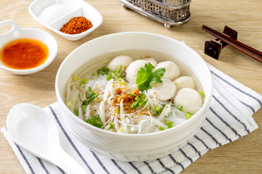 Clear noodle soup with pork meatballs in a white bowl on a wooden table.