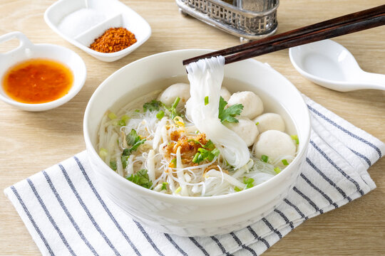 Clear noodle soup with pork meatballs in a white bowl on a wooden table.