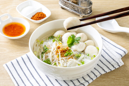 Clear noodle soup with pork meatballs in a white bowl on a wooden table.