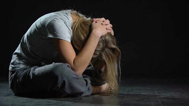 Sad woman sitting on floor in darkness with head down and hands clasped over hair, expressing emotional pain or depression