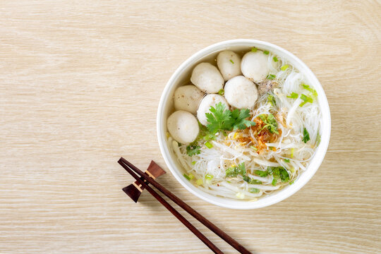 Clear noodle soup with pork meatballs in a white bowl on a wooden table.