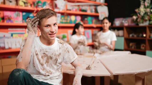 Happy student looking while put paintbrush behind ear in art lesson. Diverse student having pottery class together. high school boy smile while wearing white shirt with mud stained cloth. Edification