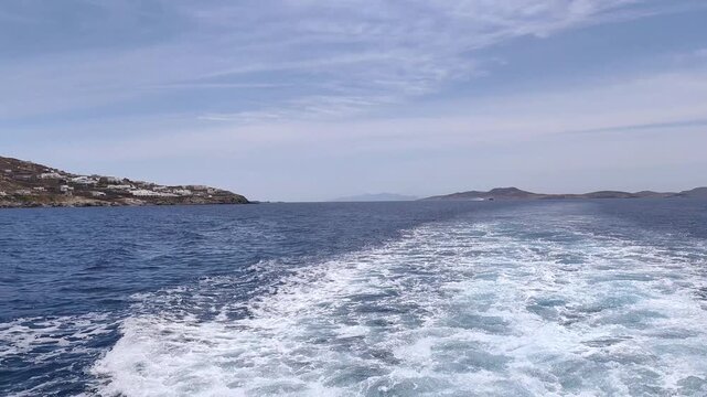 A sweeping view of a ship&rsquo;s wake as it travels away from Delos island and heads toward the island of Mykonos, cutting through the deep-blue waters of the Aegean Sea in Greece.