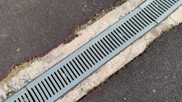 Galvanized drainage grate along stadium curb, long metal channel between asphalt and concrete, repeating slot pattern, leading