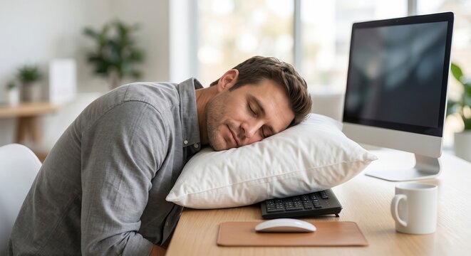 Exhausted male office worker sleeping on a white pillow at his desk next to a computer. Concept of burnout, overtime work, fatigue, and poor work-life balance in corporate environments.