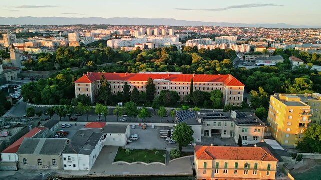 Zadar, Croatia - 05 17 2025: Angled still drone video of Zadar coastline at sunset, showing Adriatic Sea and shore on the right, city buildings on the left, Vladmir Nazor&rsquo;s Grammar School centered