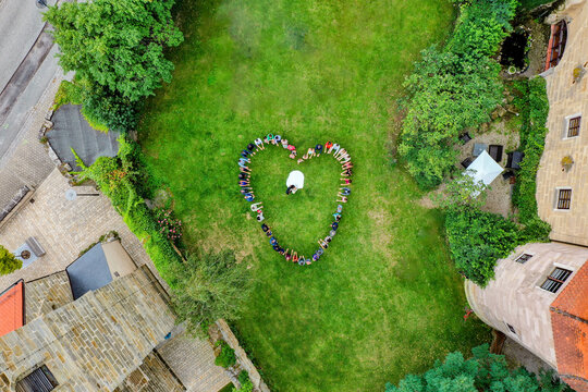 Wiesenthau, Germany - 02 May 2026: Aerial view of Schloss Wiesenthau with wedding guests forming a heart shape around a bride and groom on a green lawn.