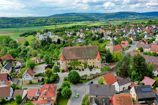 Wiesenthau, Germany - 02 May 2026: Aerial view of Schloss Wiesenthau surrounded by traditional village houses, green trees, and rolling hills under a cloudy sky.