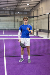Obraz premium Male athlete wearing blue top standing at net on purple padel court holding racket and ball