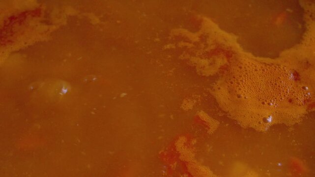 The process of preparing a home-cooked meal. A rich meat and potato soup simmers in a rustic cast-iron pot. Bubbles burst on the surface of the hot broth, steaming.