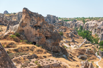 Fototapeta premium Goreme, Nevsehir, Turkey. Rock formations with carved ancient dwellings and caves, dry terrain with sparse vegetation, people walking on path, clear blue sky, bright daylight.