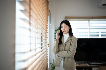Fototapeta premium Young Asian Businesswoman in blazer Talking on Smartphone by Window with Wooden Blinds in Modern Office, ideal for business communication and remote work concepts