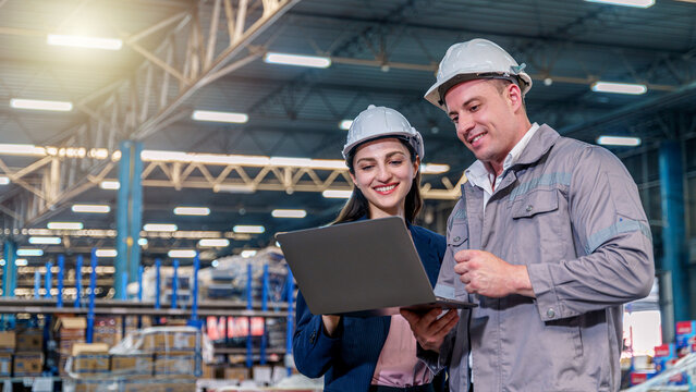 A smiling female manager in a blue blazer and a male worker in grey coveralls, both wearing white hard hats, stand inside a busy, well-lit warehouse.