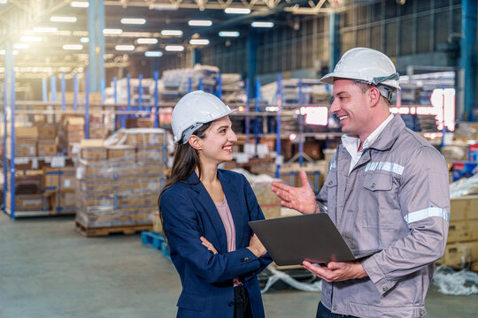 A smiling female manager in a blue blazer and a male worker in grey coveralls, both wearing white hard hats, stand inside a busy, well-lit warehouse.